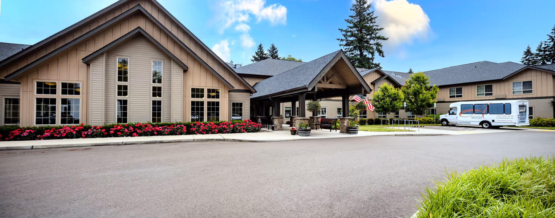 Exterior view of a senior living facility with beige siding, large windows, a covered entrance with stone pillars, American flags, landscaped flower beds, and a parked shuttle bus on a clear day.