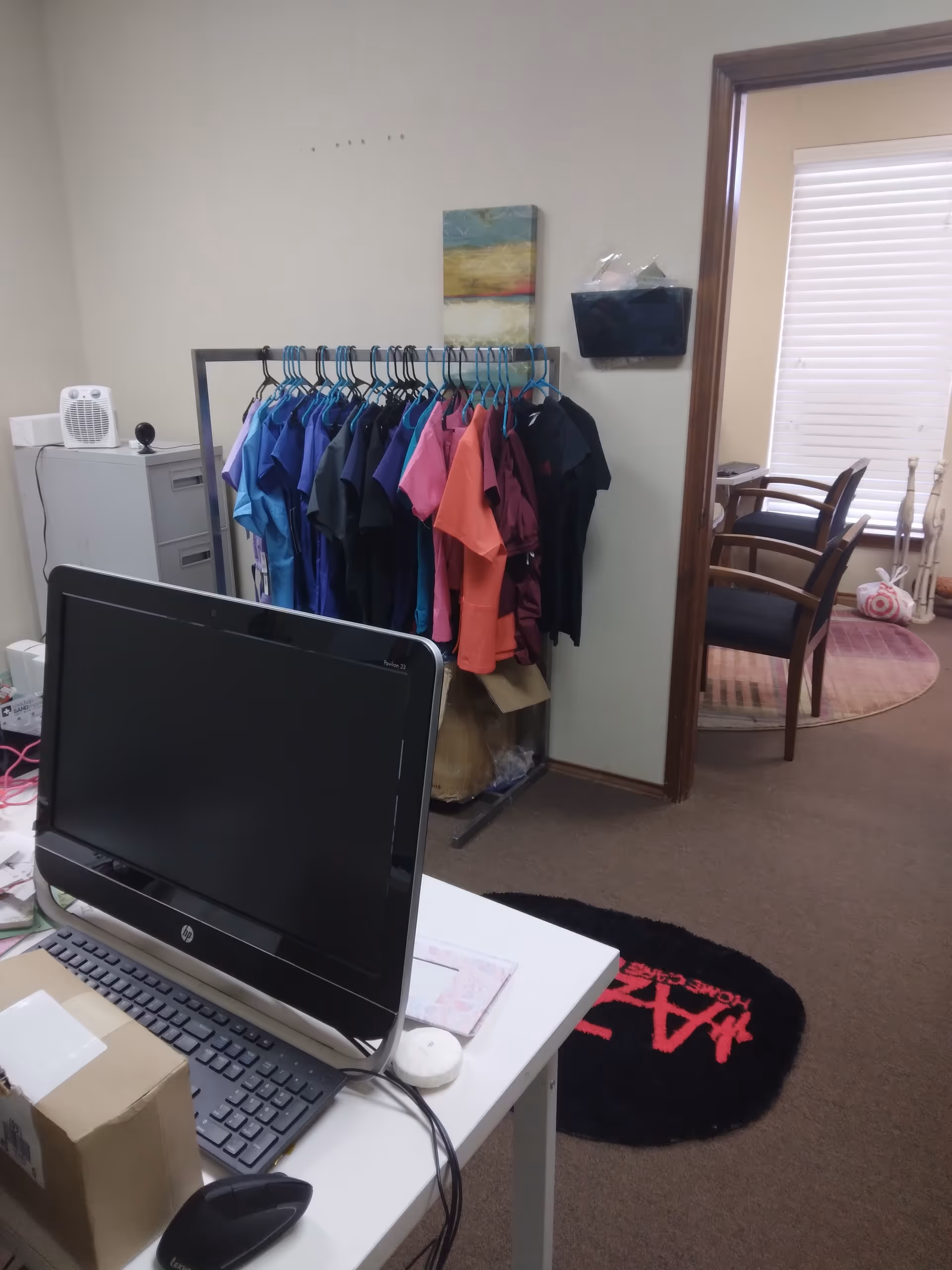 Office workspace with computer on desk, colorful scrubs on a clothing rack, doorway to seating area.