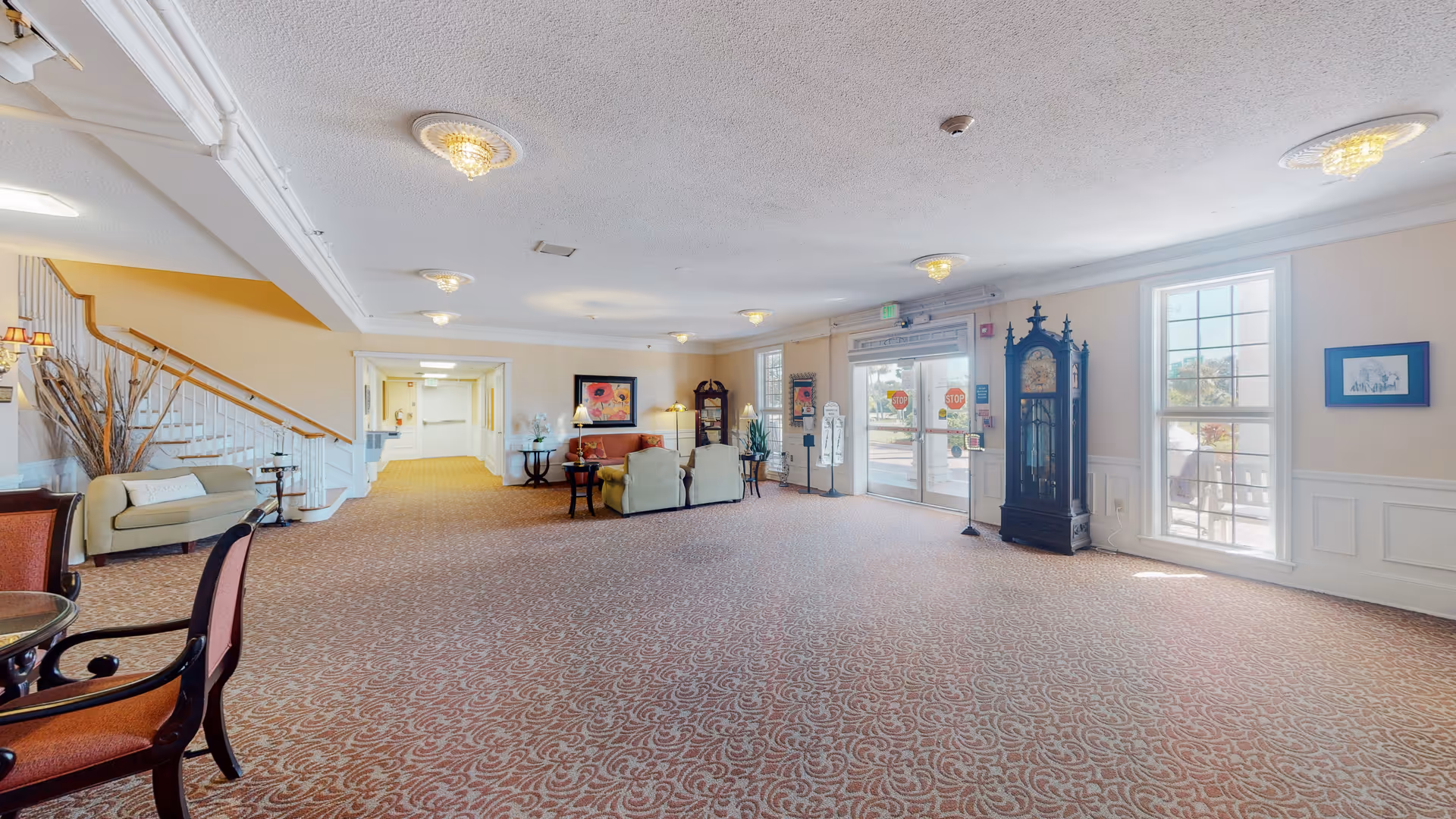 Spacious senior living lobby with seating, staircase, grand clock, and glass doors.