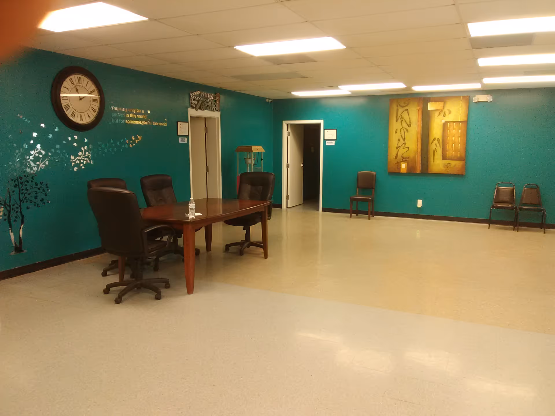 Teal-walled common room features a round clock, wall art, and a wooden table with chairs.
