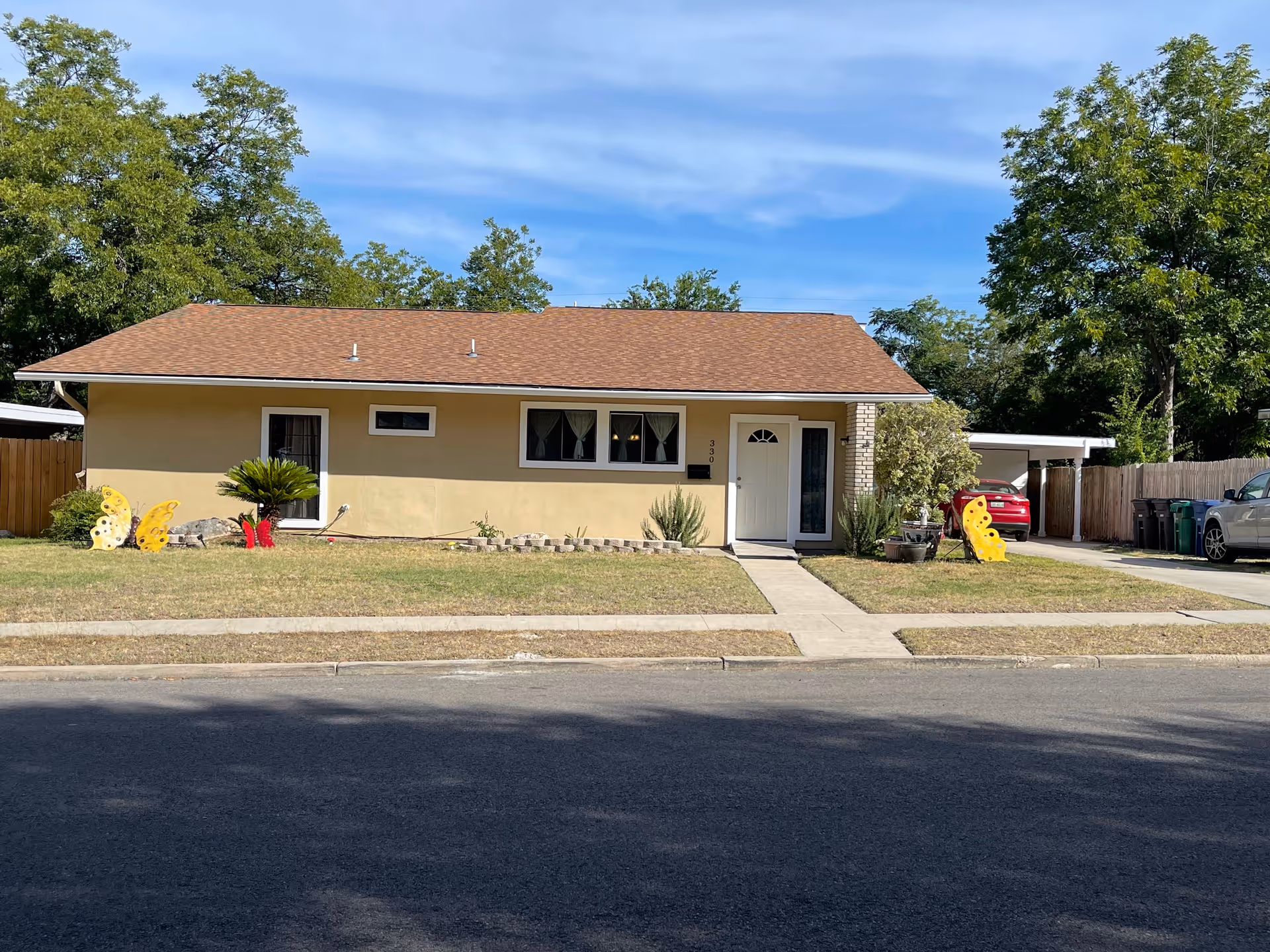 Single-story beige house with a brown roof, a white front door, and three windows with curtains. The house has a small front yard with grass, decorative butterfly yard ornaments, and a concrete walkway leading to the front door. There are trees in the background and cars parked in the driveway to the right.
