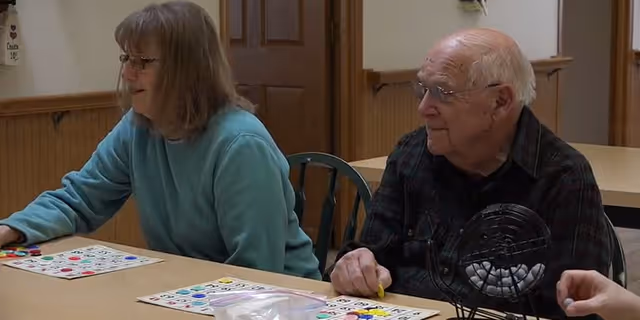 Two elderly individuals sitting at a table playing bingo. The woman on the left is wearing glasses and a light blue sweater, and the man on the right is wearing glasses and a dark plaid shirt. There is a bingo cage with balls on the table in front of them.