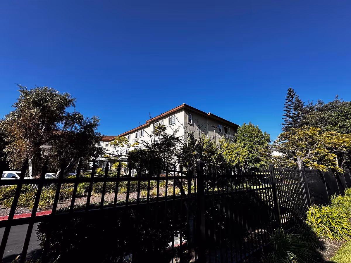 Exterior view of a multi-story residential building behind a black metal fence with trees under a clear blue sky.