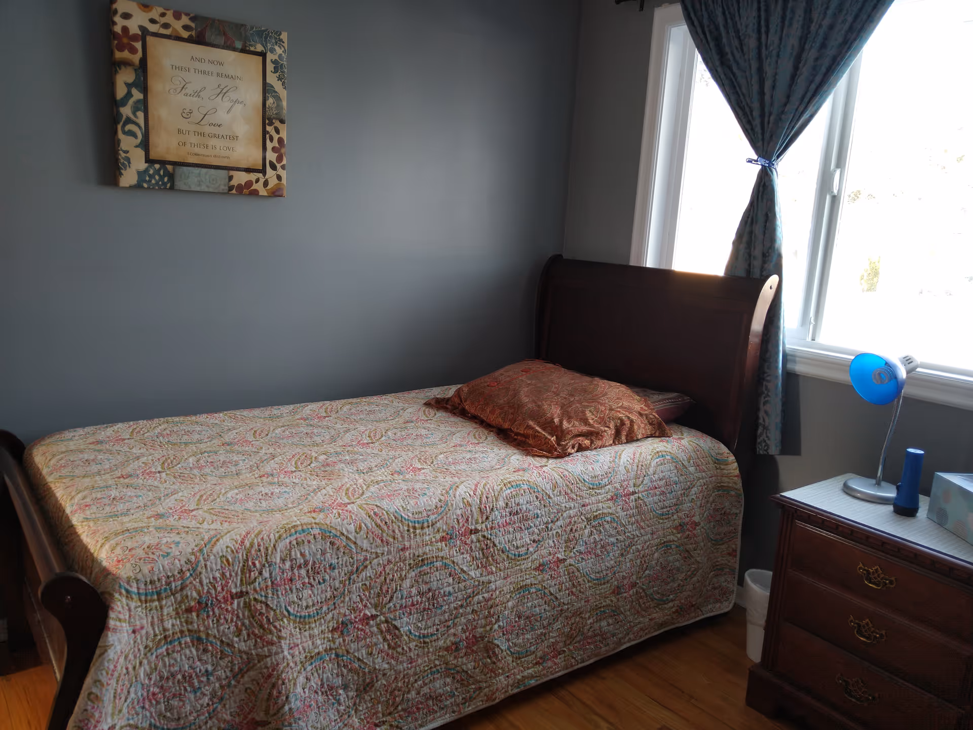 A tidy bedroom with quilted bed, dresser, lamp, and window with curtains.