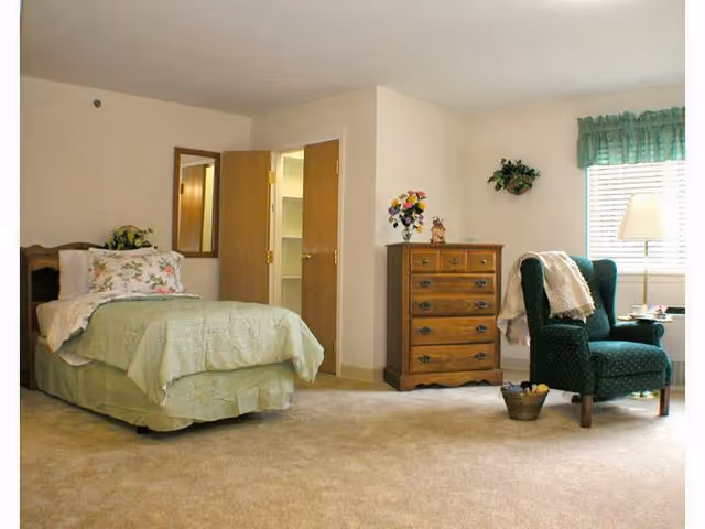 A bedroom in a senior living community with a bed, dresser, chair, and window.