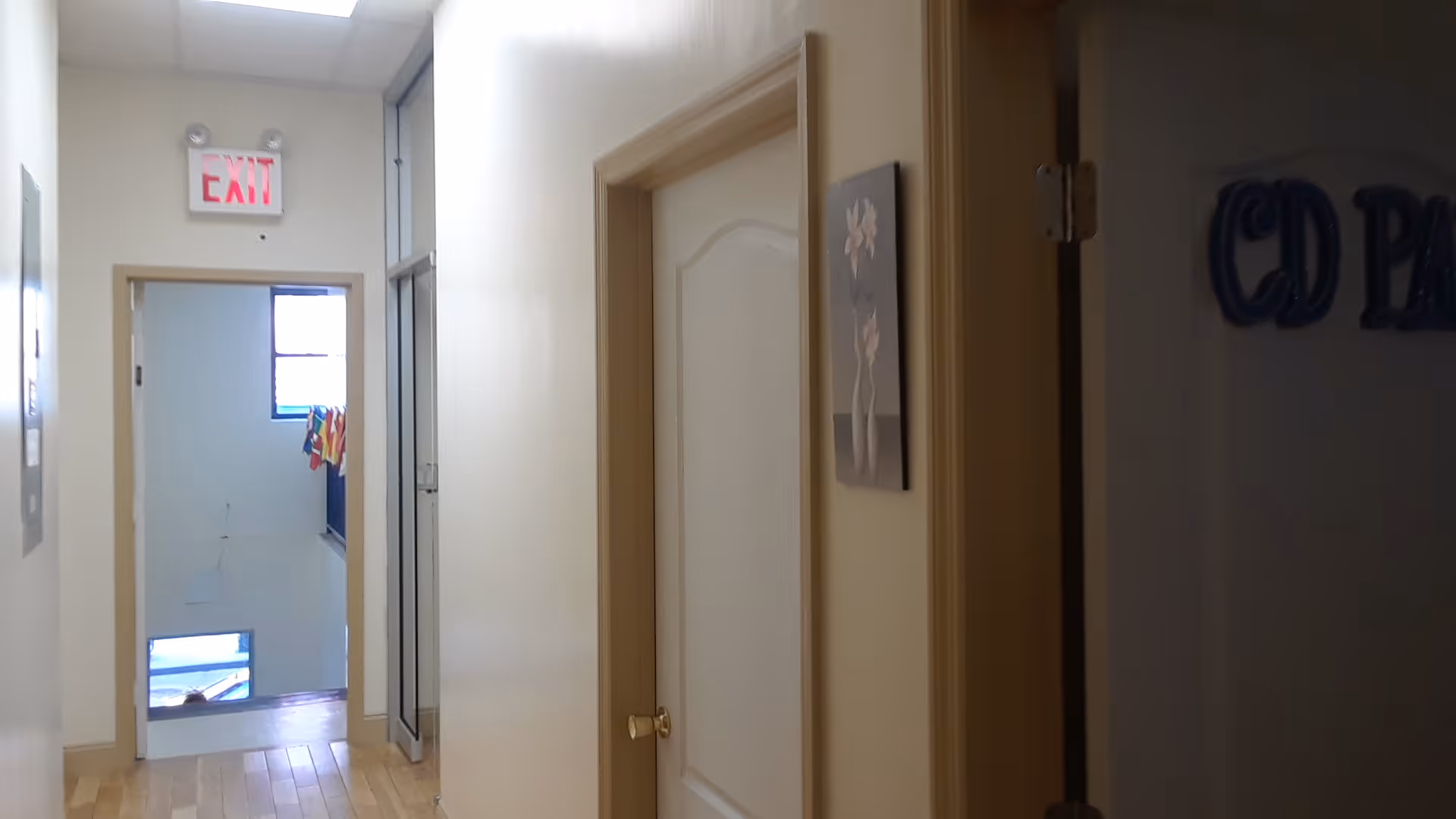 Narrow beige corridor inside a senior living facility with doors, artwork, and an exit sign.