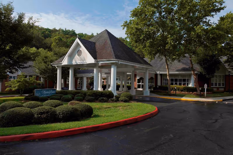 Covered porte-cochère entrance of a brick senior living building with manicured shrubs and a curved driveway.