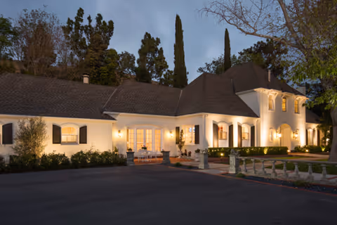 Exterior view of a large, elegant building at dusk with warm lights illuminating the facade, surrounded by trees and a paved driveway in front.