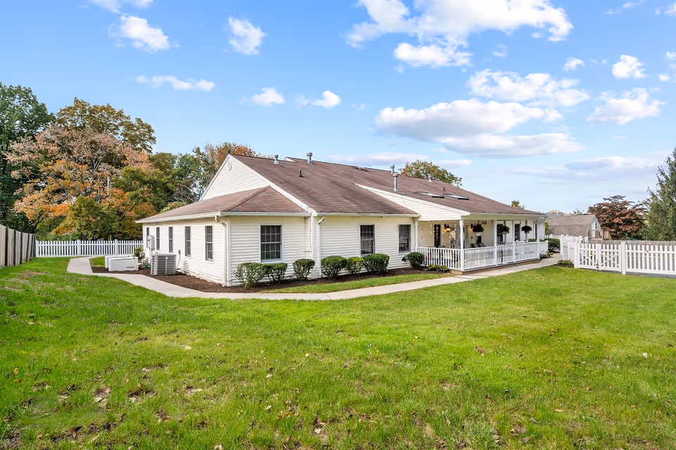 Single-story memory care facility exterior with a white facade, green lawn, and wraparound porch.
