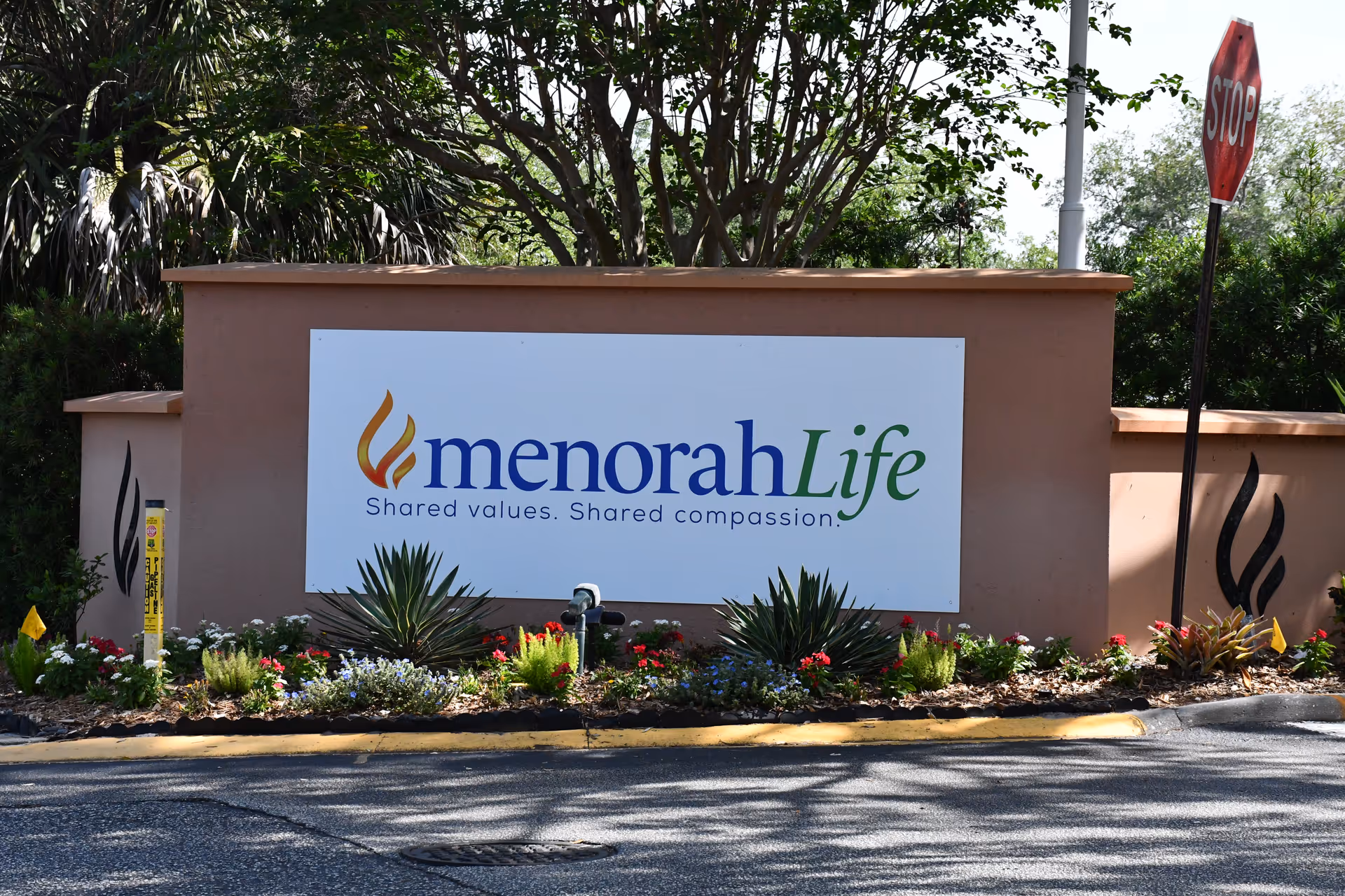Front entrance sign for menorahLife with landscaping and a nearby stop sign.