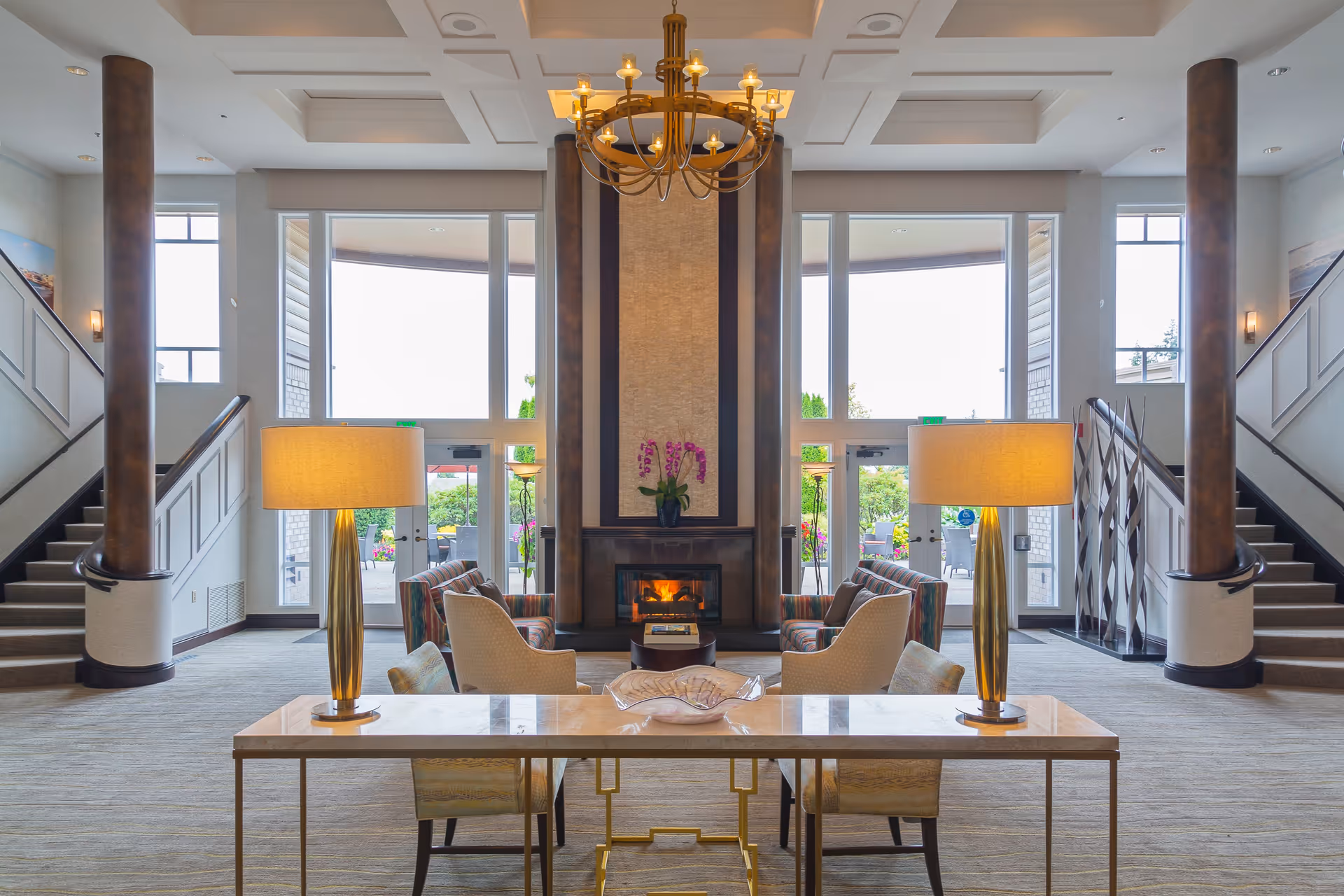 A spacious and elegant living room area with a central fireplace flanked by two tall pillars. There are two sets of stairs on either side leading to an upper level. The room features large windows and glass doors that open to an outdoor patio area. Two table lamps with golden bases sit on a marble-topped table in the foreground, with comfortable armchairs arranged around the fireplace. A chandelier hangs from the ceiling, adding a warm ambiance to the space.