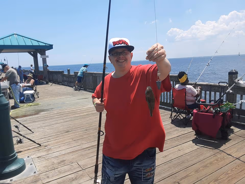 Smiling man in a red shirt holds a fish on a wooden pier by the ocean.