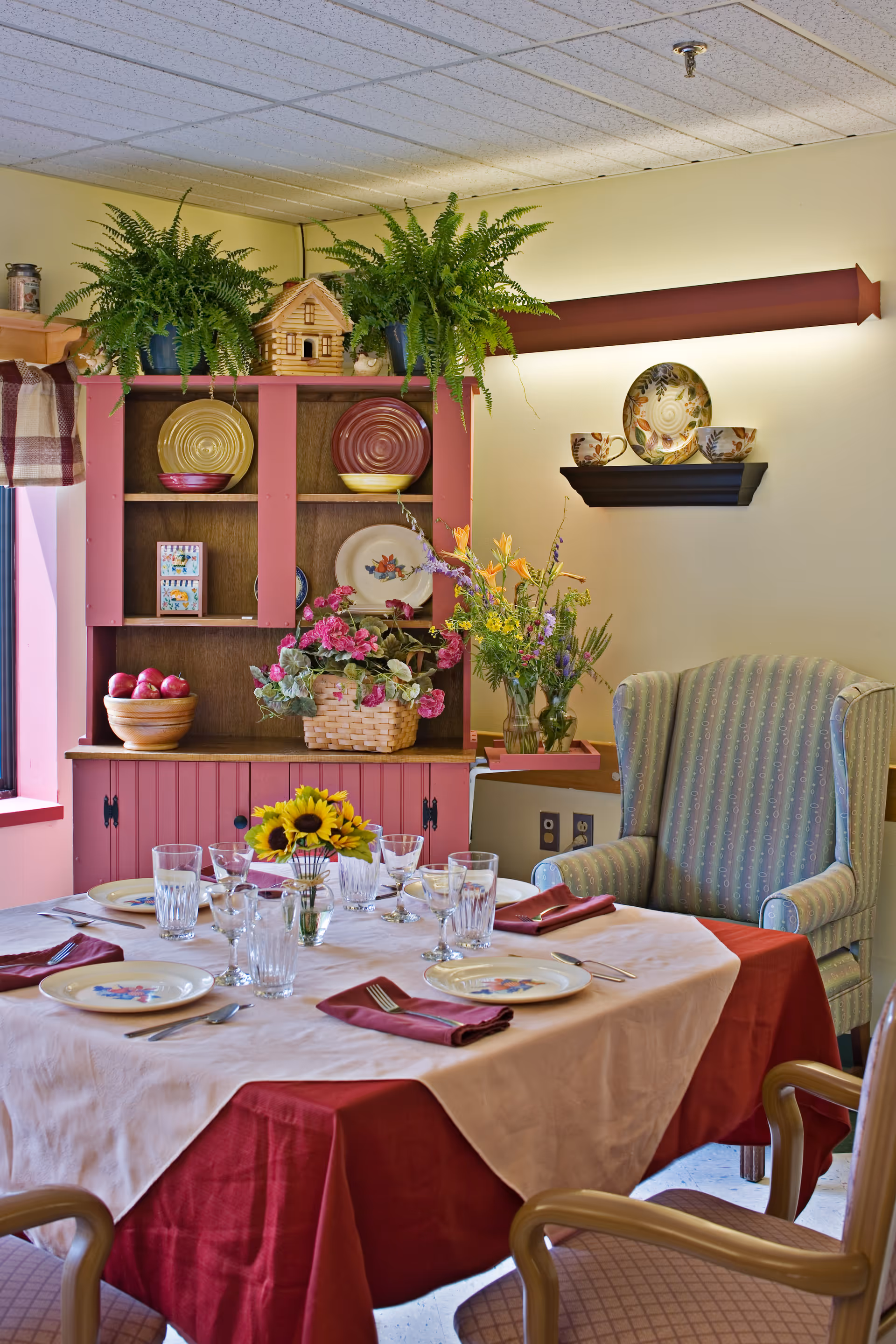 Dining table set for a meal with plates, glasses and a sunflower centerpiece in a cozy room with a pink hutch and an upholstered armchair.