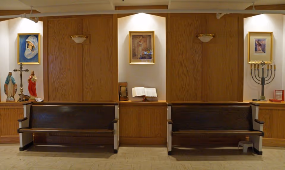 Interior view of a small chapel or prayer room with two wooden pews facing forward. The walls behind the pews have religious statues, a cross, a menorah, framed religious artwork, and an open Bible displayed on a wooden ledge. The lighting is warm and soft.