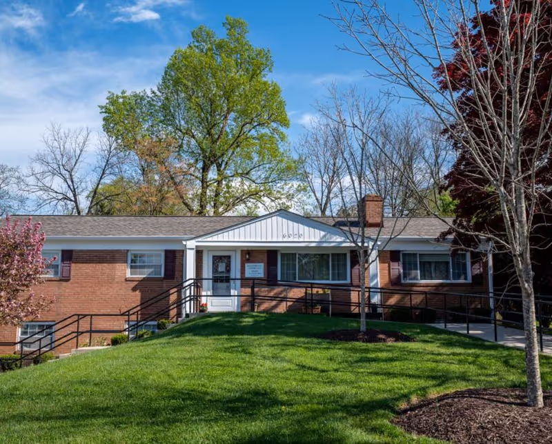 Brick, single-story facility front with ramp, green lawn, and tall trees.