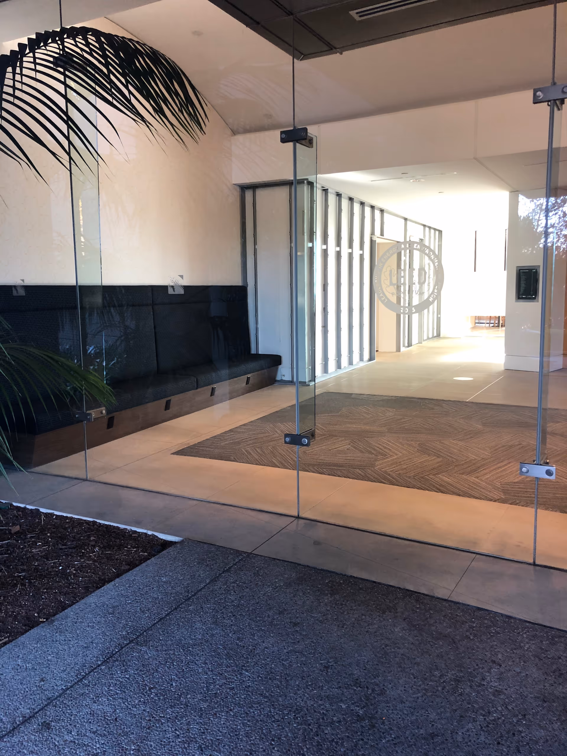 Glass entrance to a lobby with a black bench and a potted palm inside