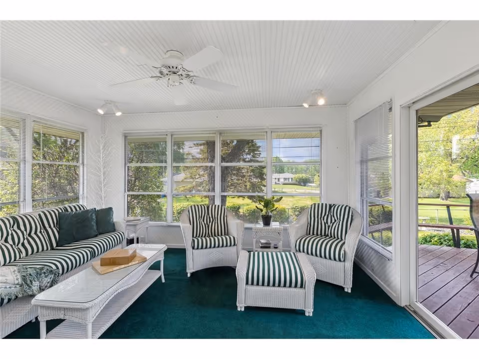 Bright sunroom with white wicker seating and large windows facing a green yard.