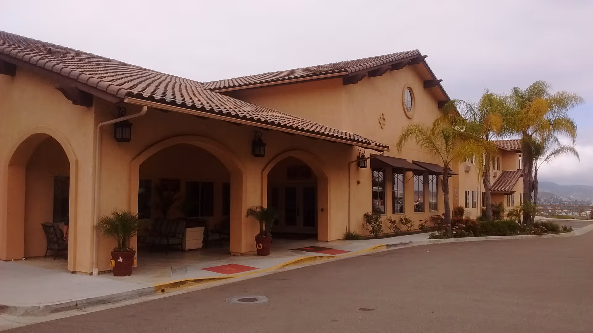Front exterior of Lantern Crest Senior Living with arches, palm trees, and a walkway.