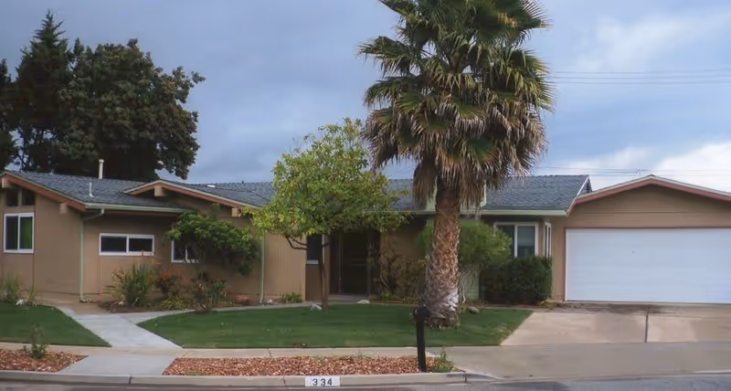 Single-story residential building with a front lawn, a palm tree, and a driveway leading to a two-car garage under a cloudy sky.