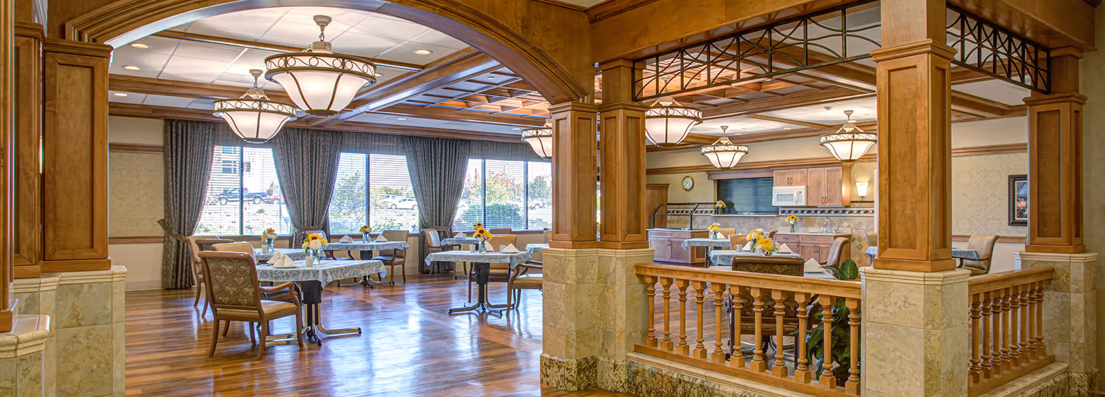 A spacious dining room in a senior living facility with wooden floors, multiple tables covered with patterned tablecloths, chairs, and floral centerpieces. Large windows with curtains allow natural light to fill the room. The ceiling features decorative wooden beams and elegant hanging light fixtures. The room is divided by wooden columns and a half-wall with balusters.
