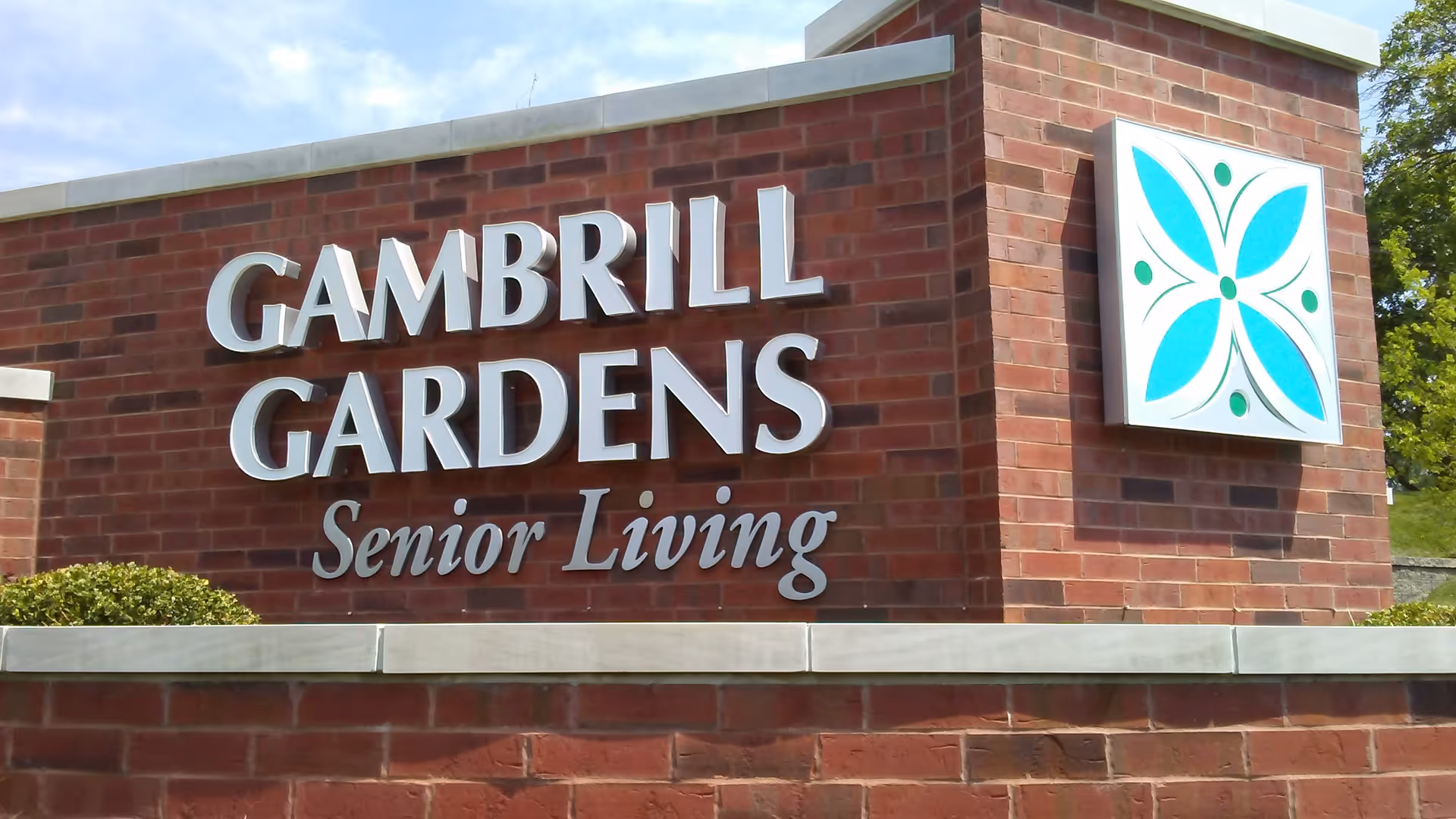 A brick sign for Gambrill Gardens Senior Living with white raised letters and a decorative blue and green floral logo on the right side.