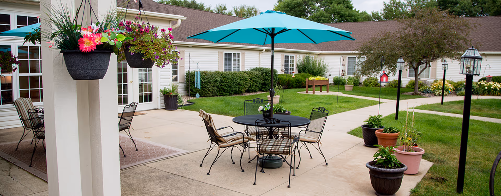 Outdoor patio area at Bickford of Fort Dodge with a round black metal table and four chairs, one with a cushion, under a large blue umbrella. There are hanging flower pots with colorful flowers on a white pillar, several potted plants on the ground, and a well-maintained lawn with bushes and trees in the background. The building has white siding and multiple windows and doors.