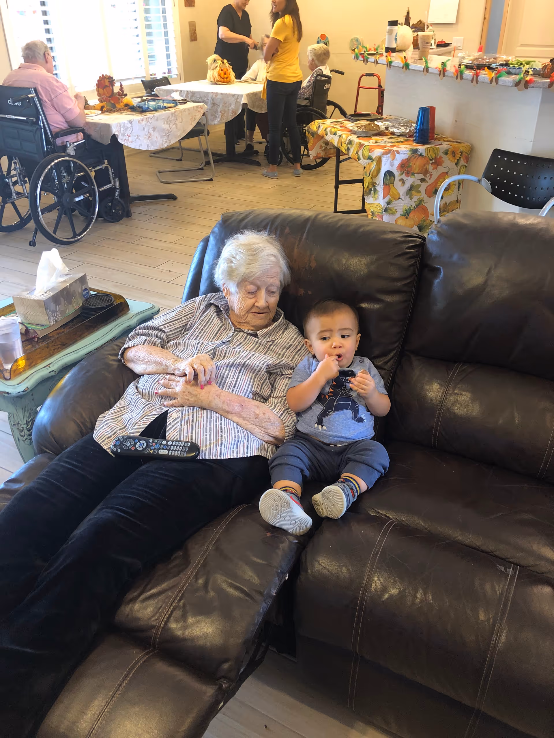 An elderly woman sits on a leather couch with a toddler in a bright living room.