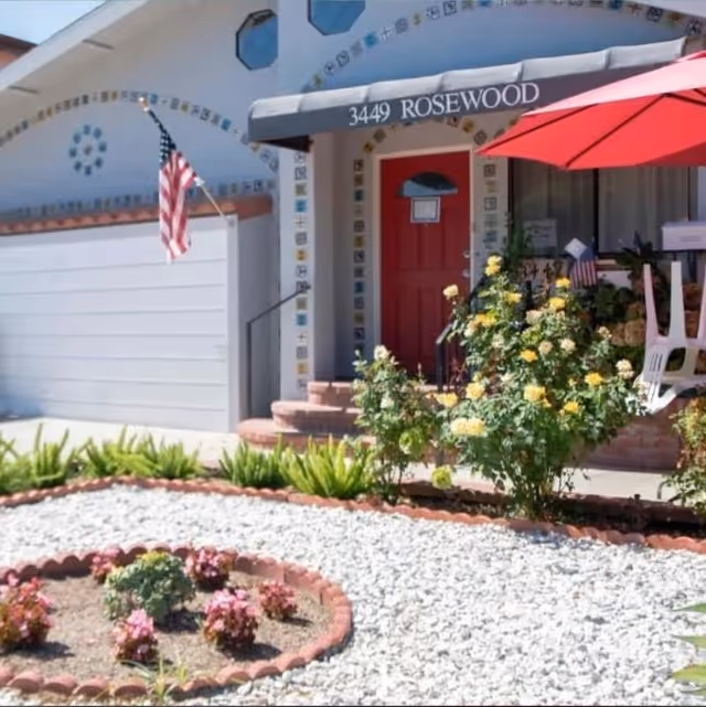 Front entrance of a house with a red door under an awning reading '3449 ROSEWOOD', an American flag, gravel landscaping, and flowering bushes.