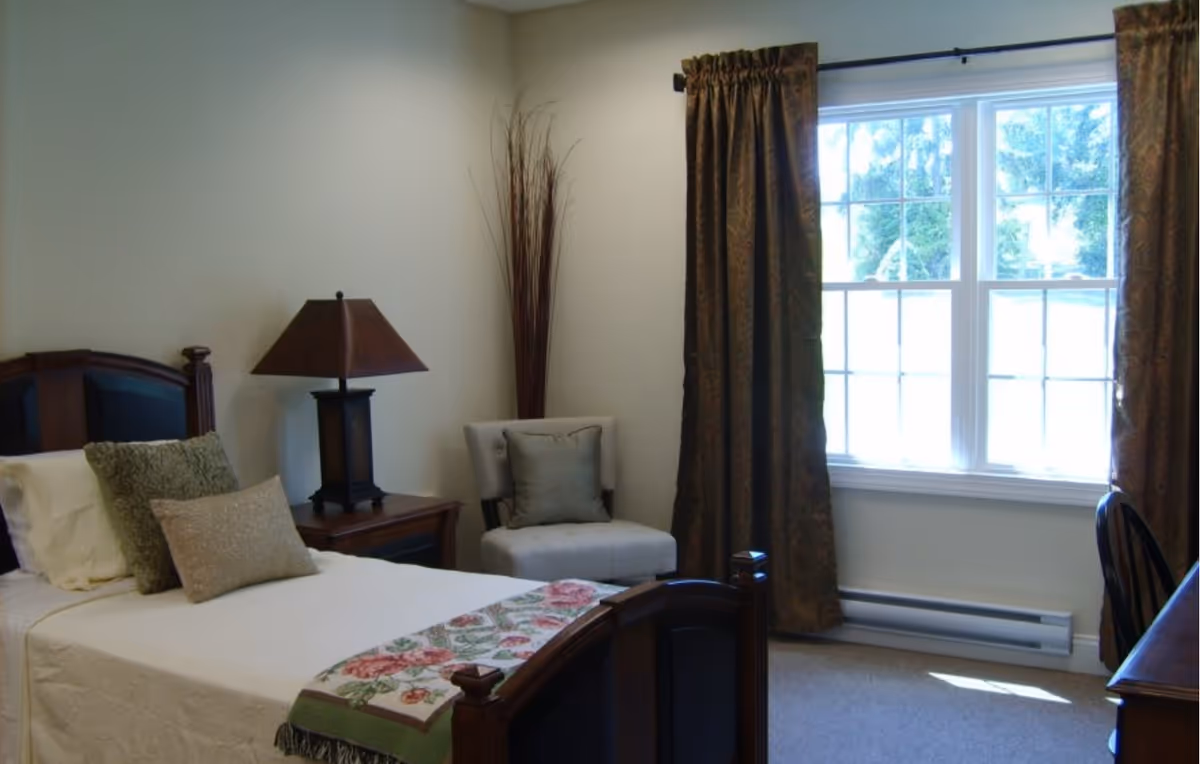 A furnished bedroom with a single bed, nightstand and lamp, an upholstered chair, and a window with curtains.