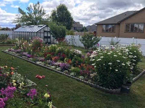 A well-maintained garden with various colorful flowers and plants bordered by a low stone edging. In the background, there is a white fence, a greenhouse, and houses under a partly cloudy sky.