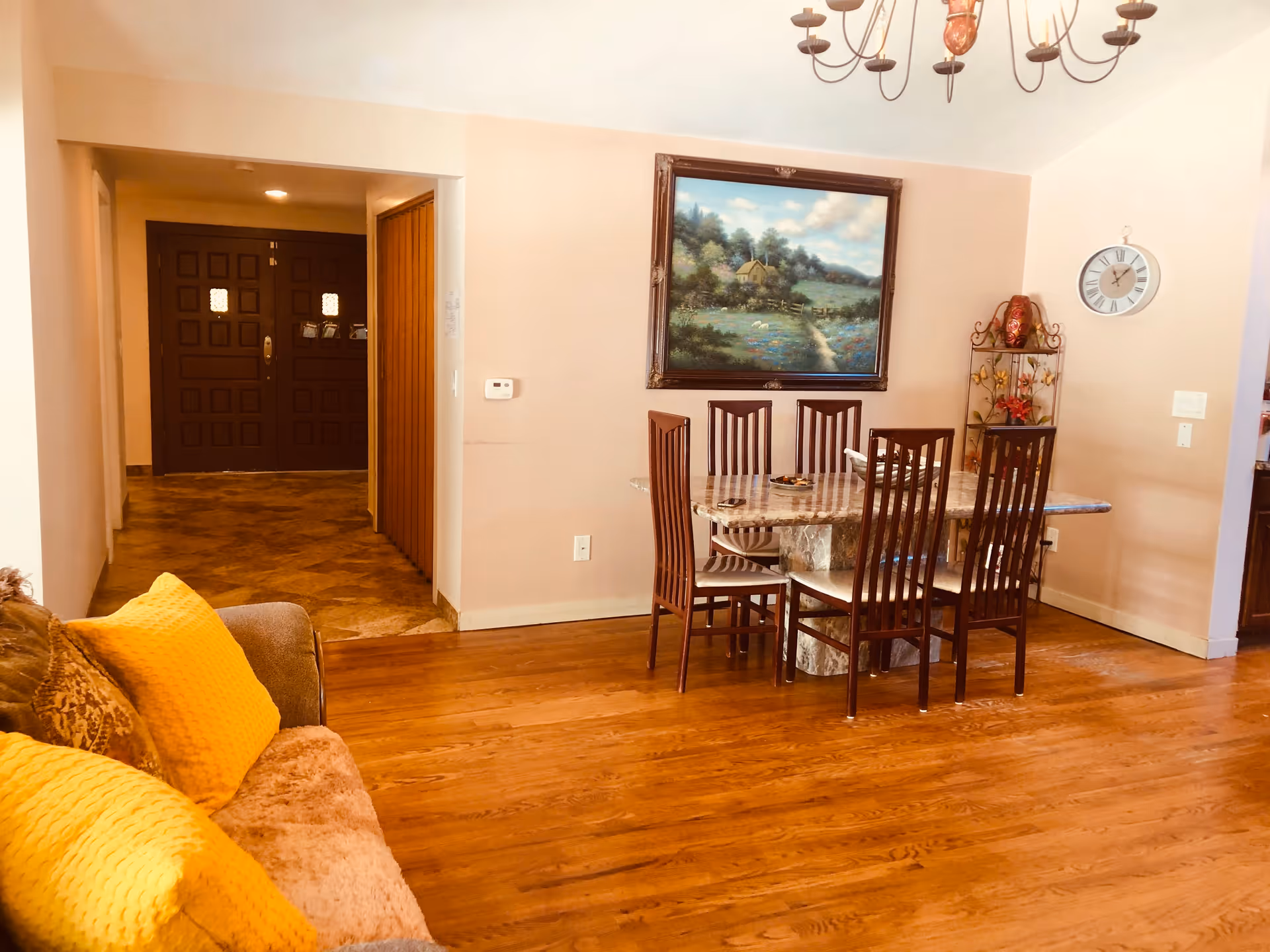 Interior view of a senior living facility showing a dining area with a rectangular marble table surrounded by six wooden chairs. A large framed painting of a countryside scene hangs on the wall above the table. To the right, there is a decorative metal shelf with a vase and flowers, and a round wall clock. On the left side, part of a brown couch with yellow cushions is visible. The floor is wooden, and in the background, there is a hallway leading to a dark wooden double door.