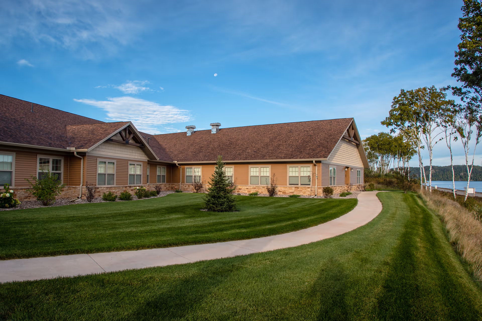 Exterior view of a single-story senior living facility building with brown siding and a brown shingled roof, surrounded by a well-maintained green lawn, a curved concrete walkway, and trees near a body of water under a blue sky with some clouds.