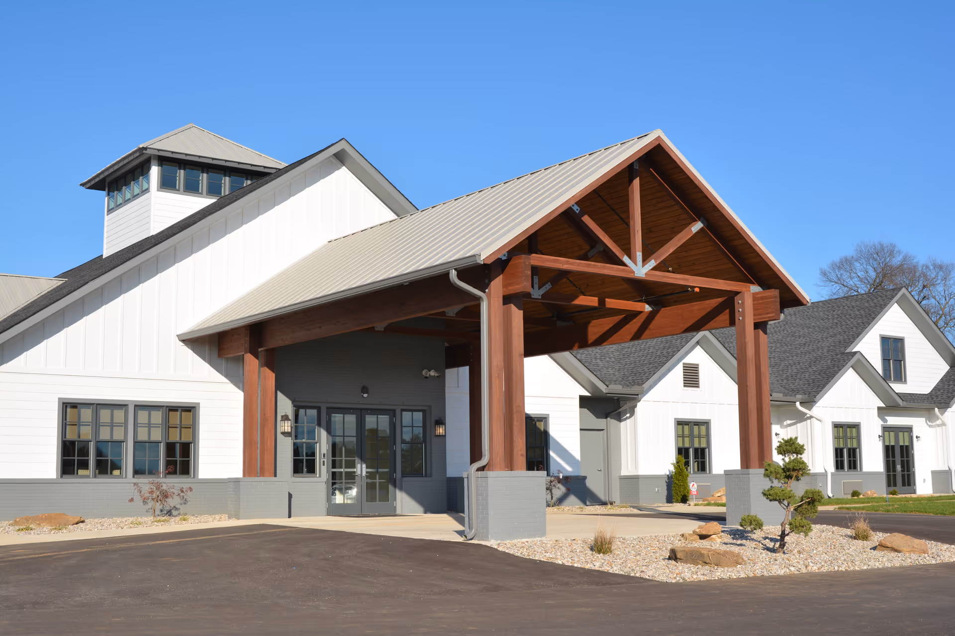 Exterior view of Lewis Manor Assisted Living facility showing a modern building with white siding, gray roof, large wooden entrance canopy, and multiple windows under a clear blue sky.
