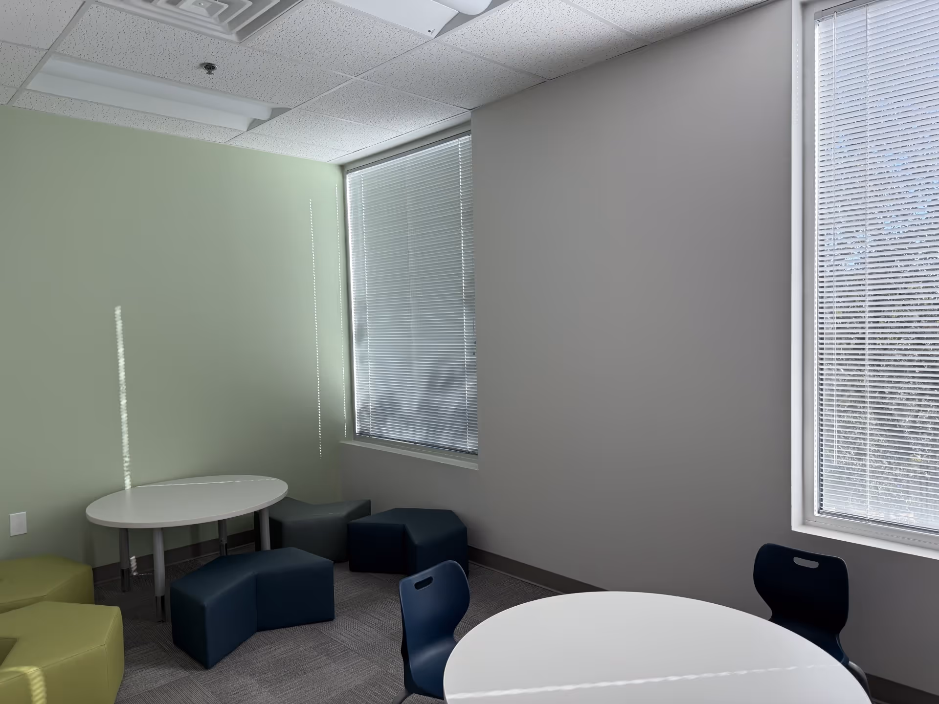 Bright, sparse lounge area with round tables, cube stools, and blinds-covered windows.