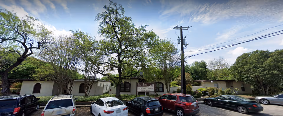 Exterior view of San Jose Nursing Center showing a single-story building partially obscured by trees and bushes. Several parked cars are visible in front of the building under a partly cloudy sky.