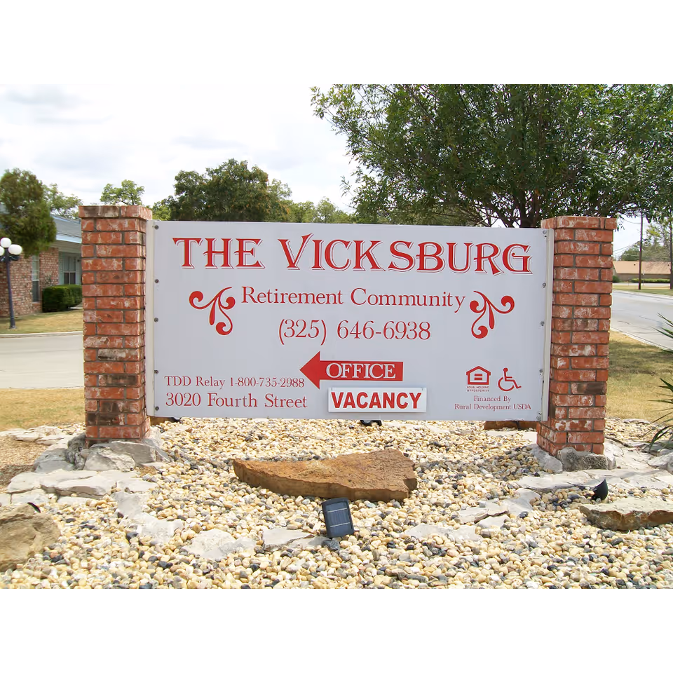 A freestanding sign between brick pillars reading "The Vicksburg Retirement Community" with contact details and a "VACANCY" notice in a rock landscaped front area.