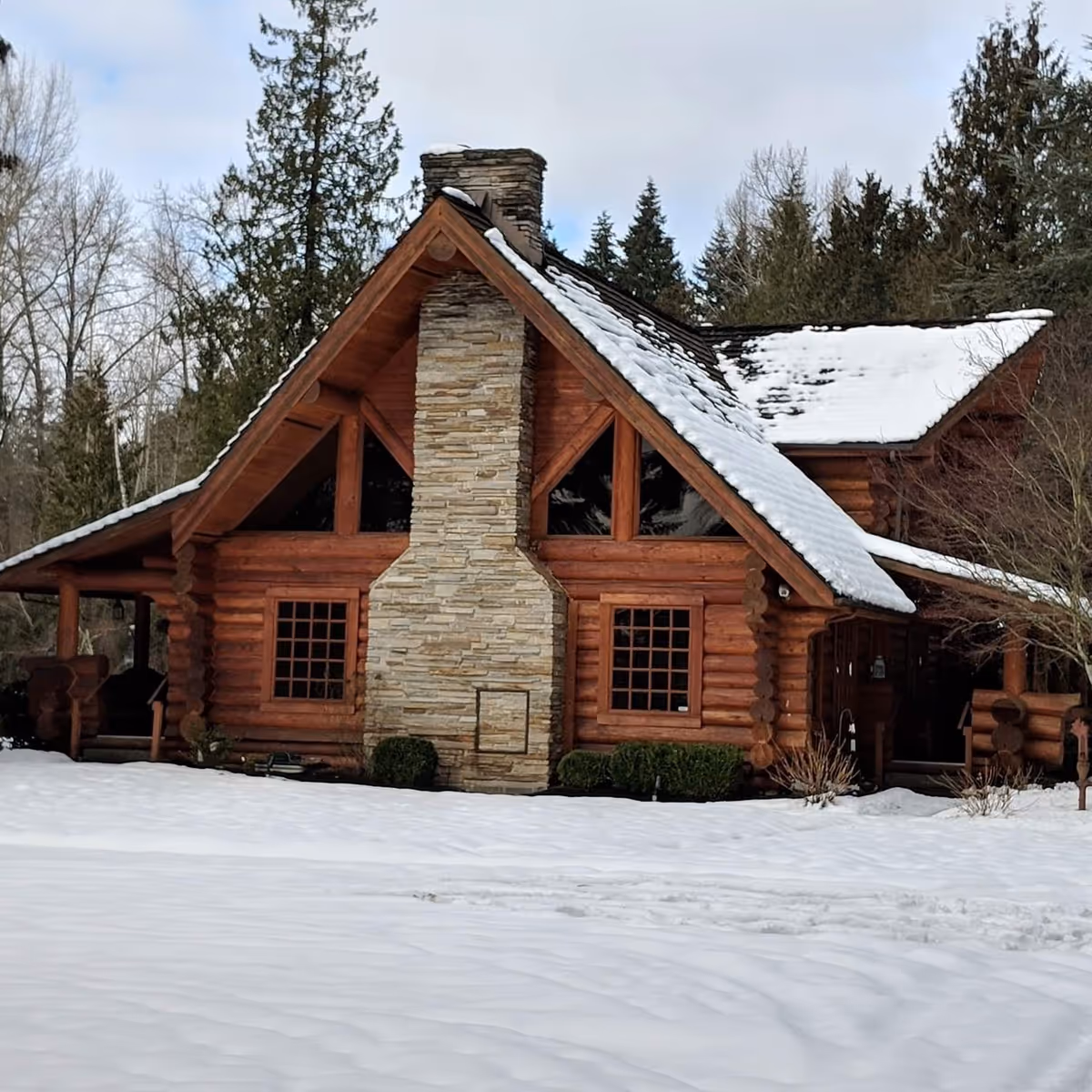 Snow-covered log cabin exterior with a stone chimney and surrounding evergreen trees.