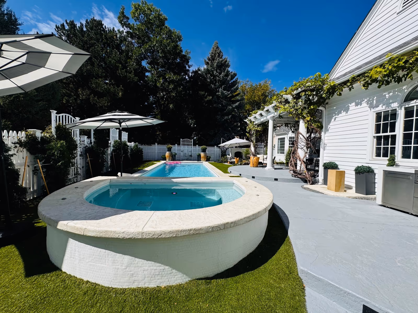 Sunny outdoor pool and hot tub area beside a white building with umbrellas and greenery.