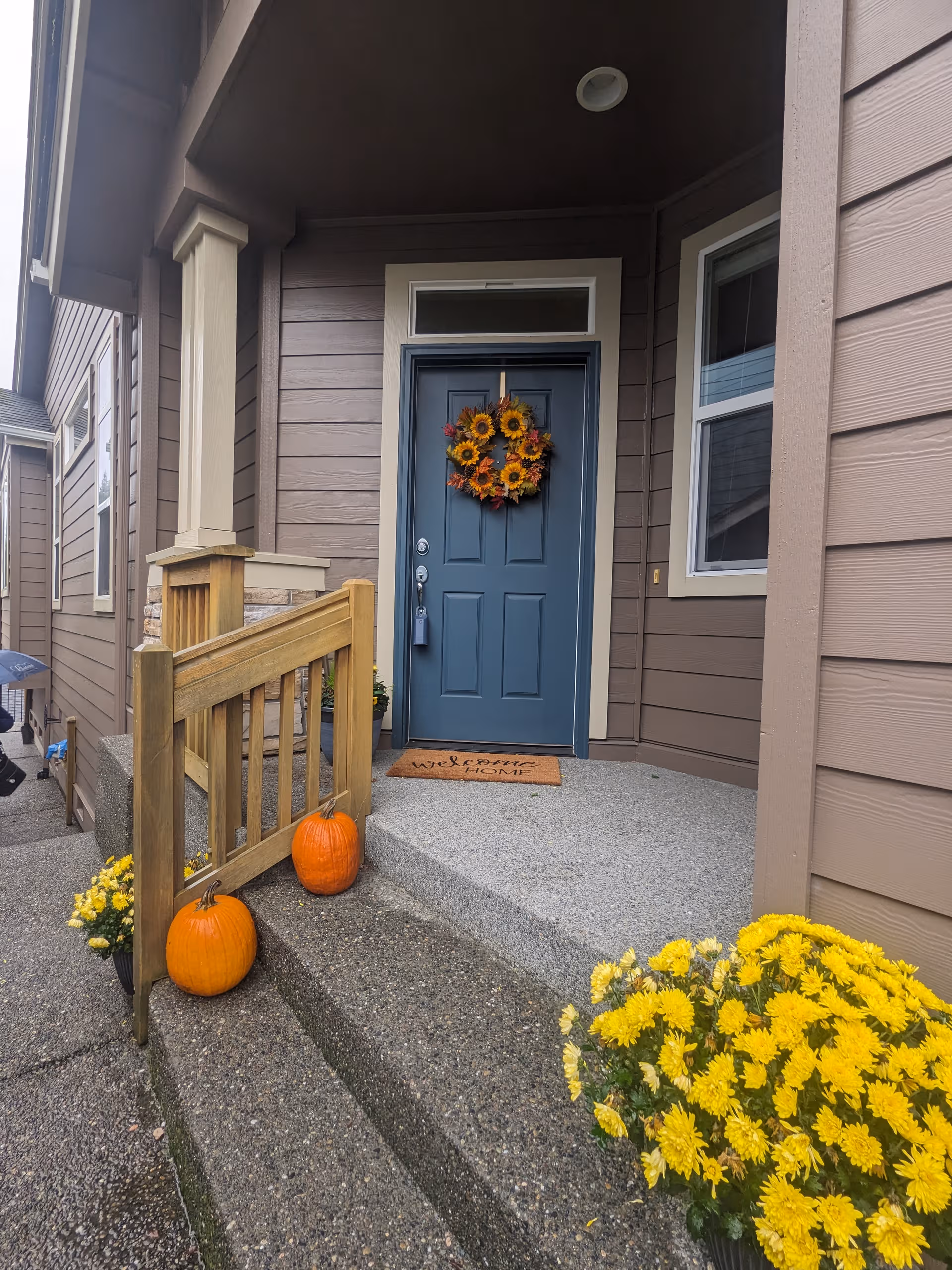 Front entrance of a brown building with a blue door, wreath, pumpkins, and yellow mums.