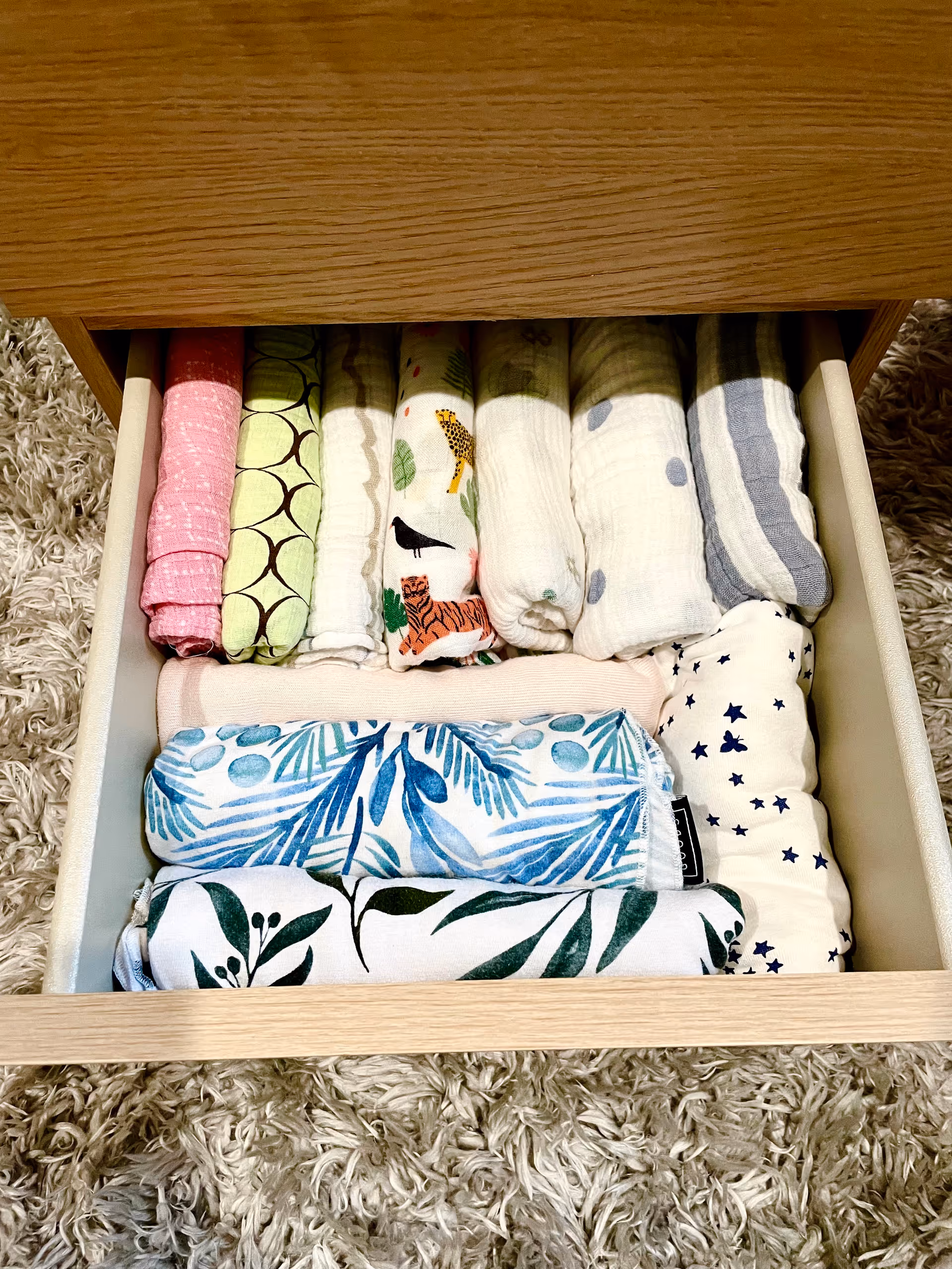 Open wooden drawer filled with folded colorful fabric and towels on a shag rug.