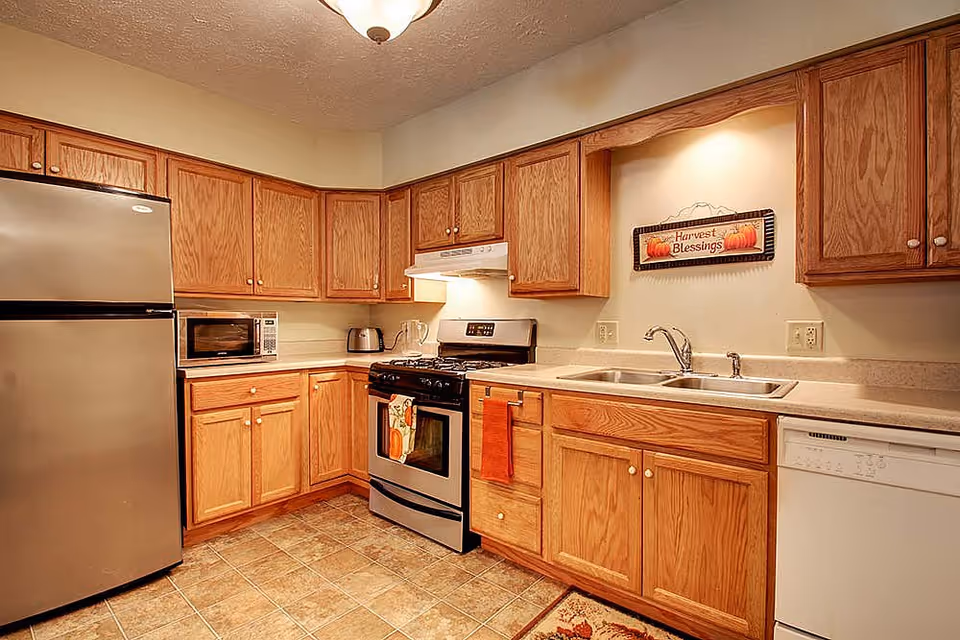 A kitchen with wooden cabinets, a stainless steel refrigerator, a microwave, a toaster, a gas stove with an oven, a double sink, and a dishwasher. There is a decorative sign on the wall above the sink that reads 'Harvest Blessings'.