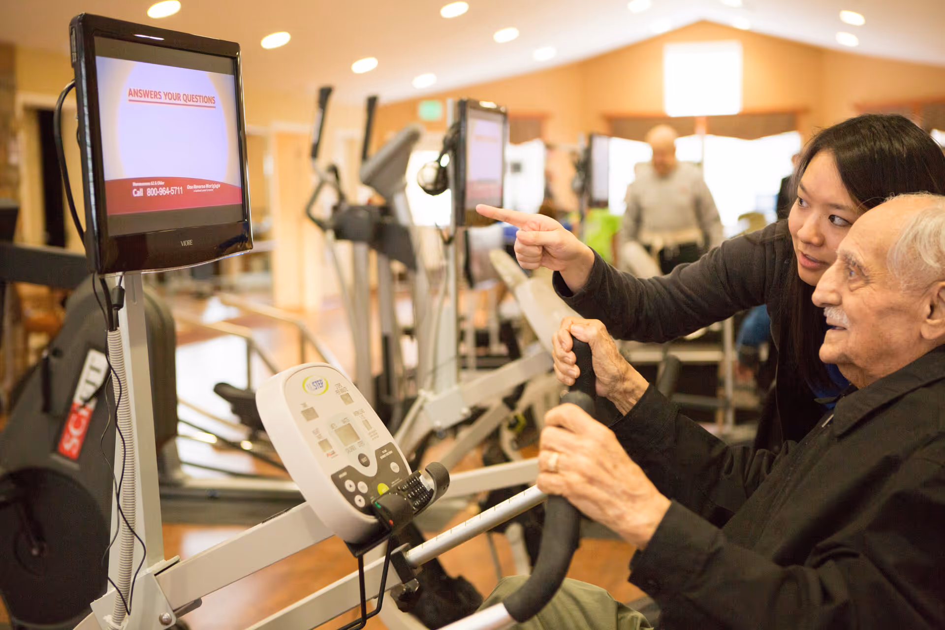 An elderly man using a stationary exercise bike in a fitness room, guided by a younger woman who is pointing at a screen attached to the bike. Other exercise equipment is visible in the background.