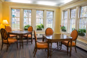 A bright dining area with multiple wooden tables and cushioned chairs arranged near large windows that let in natural light. The room has light yellow walls, a striped carpet, and several potted plants on the windowsills.