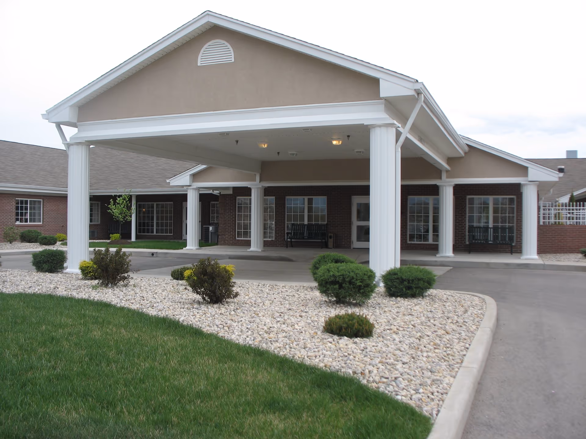 Front entrance of a single-story building with a covered driveway supported by white columns, surrounded by landscaped bushes and a lawn.