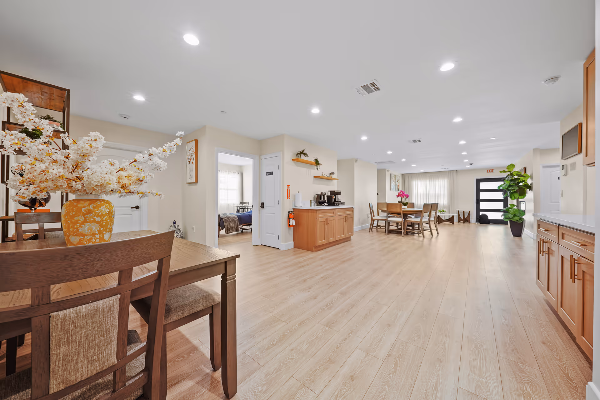 Open, well-lit senior living common area with dining tables and kitchen cabinets.