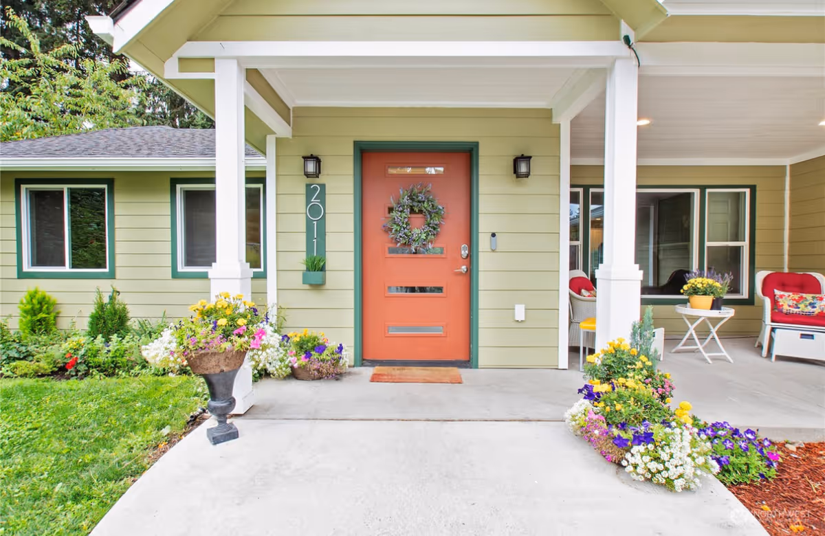 A green-painted building entrance with coral door, porch seating, and colorful flowers.