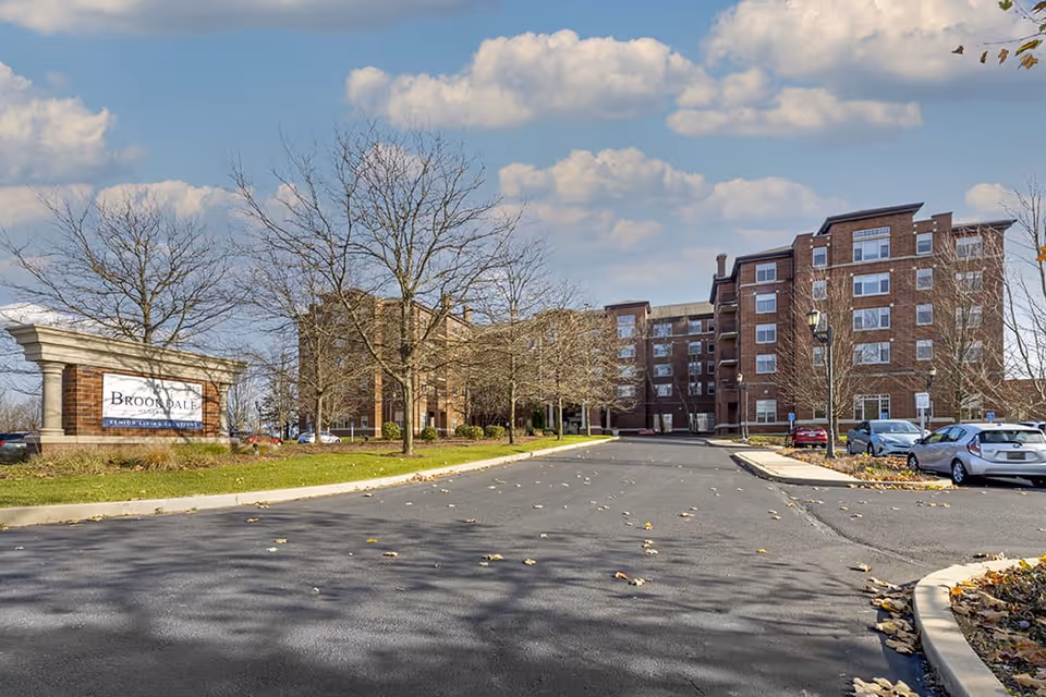 Exterior view of Brookdale Mt. Lebanon senior living facility showing a multi-story brick building with several windows, a parking lot with parked cars, leafless trees, and a sign on the left side that reads 'Brookdale Senior Living Solutions' under a partly cloudy sky.