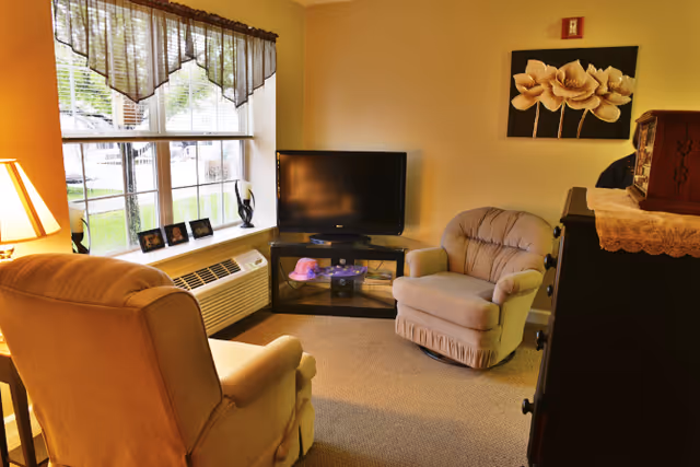 Cozy sitting area with two upholstered armchairs facing a flat-screen TV next to a large window and dresser.