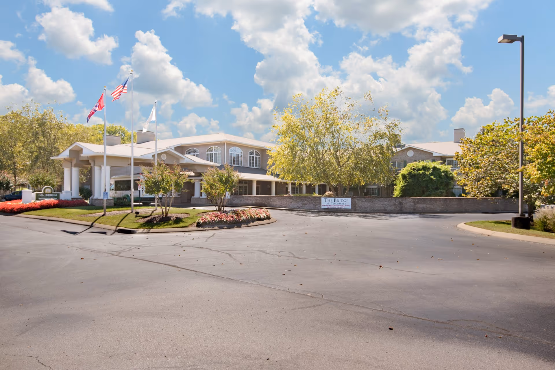 The front exterior of a senior living facility with a porte-cochère, flagpoles, circular driveway, and landscaped flower beds under a partly cloudy sky.