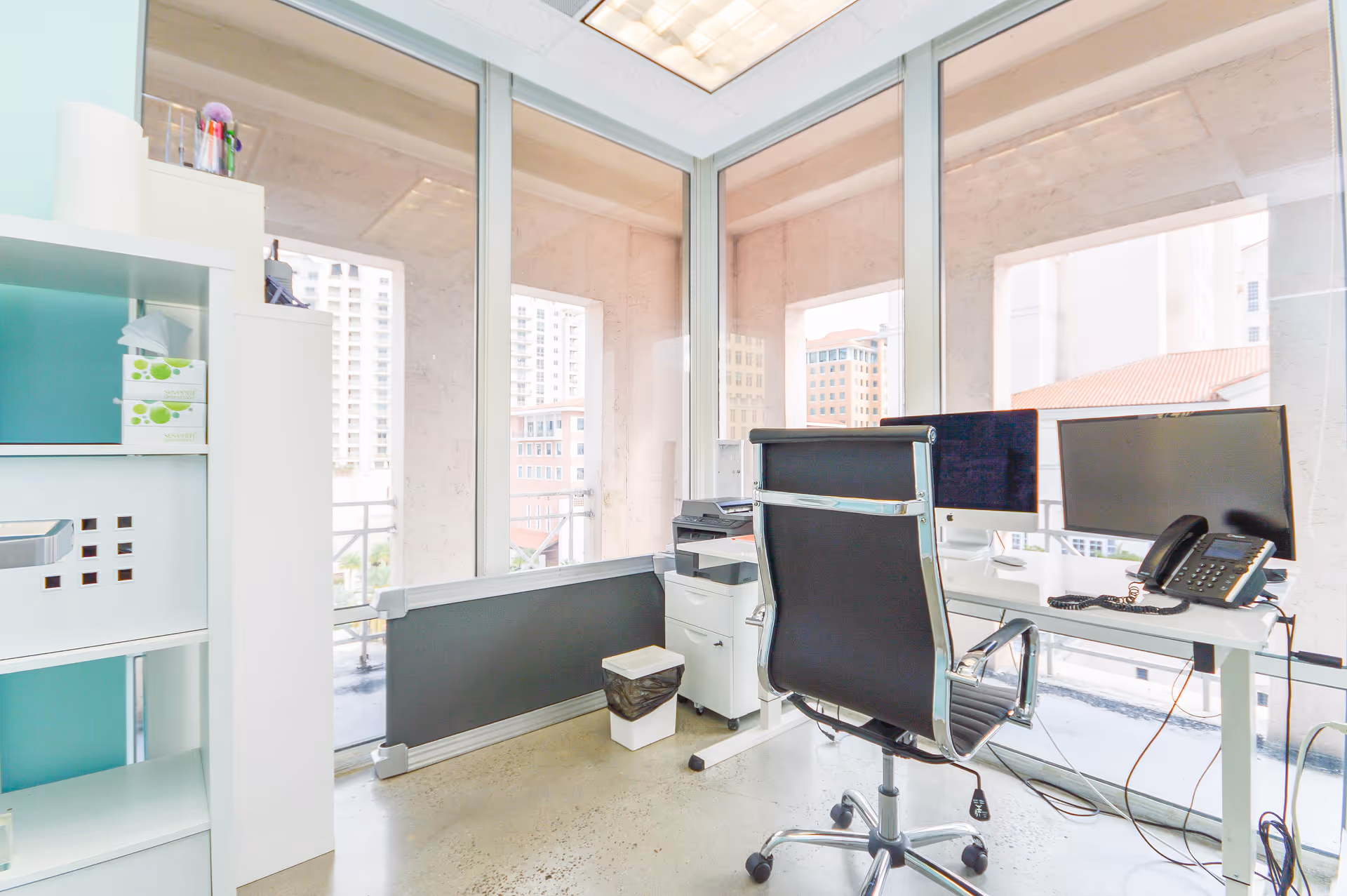 Bright modern office with dual monitors, desk, chair, shelves, and city skyline outside.